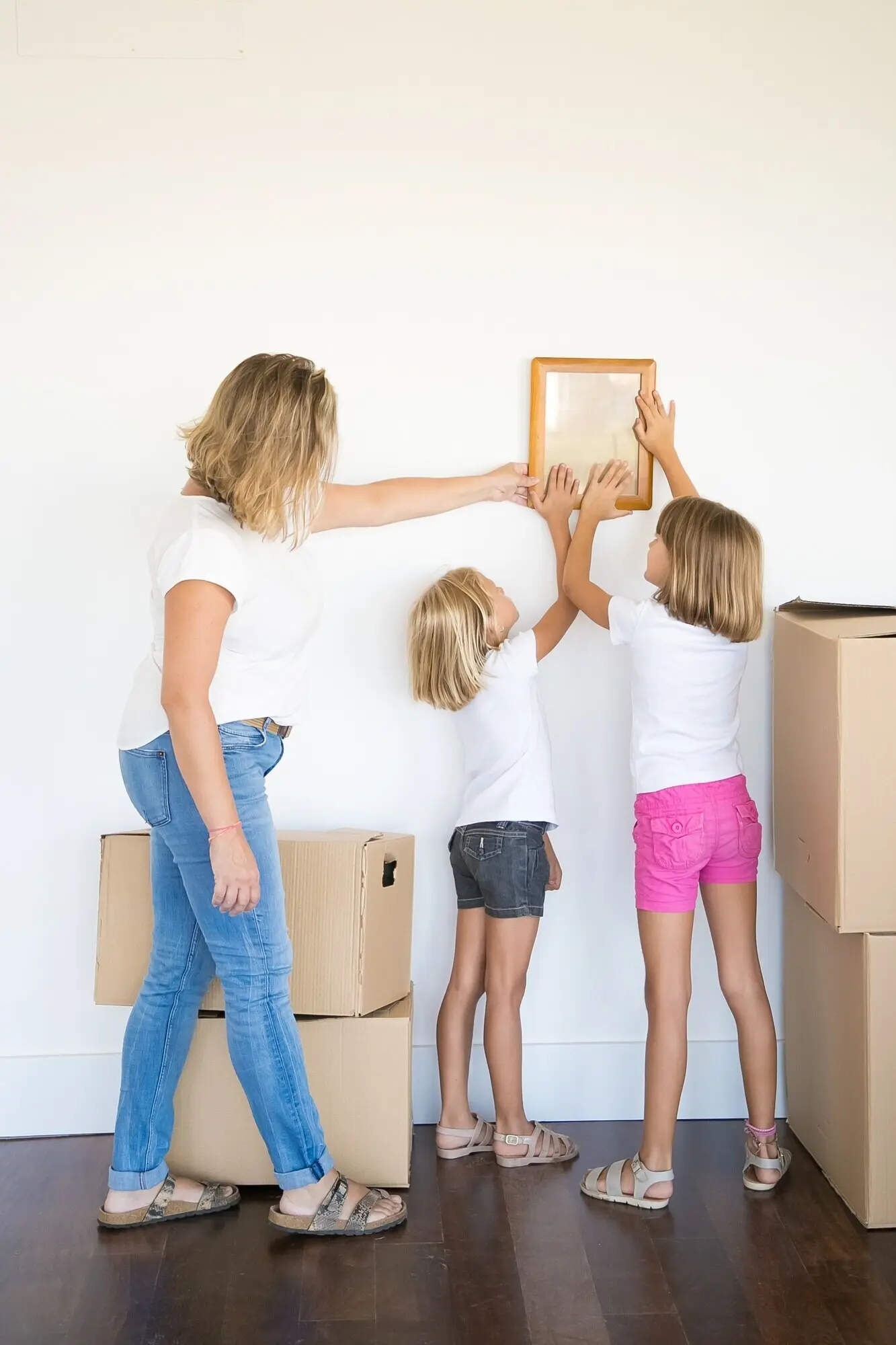 Мама и две девочки вешают пустую фоторамку на белую стену.